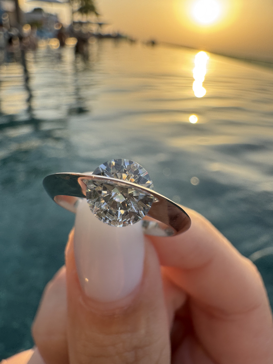 Primo piano dell’anello Giroluce tenuto tra le dita, diamante che ruota riflettendo la luce dorata del tramonto sul mare