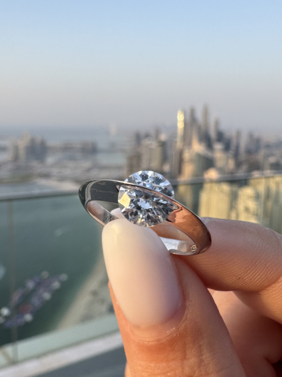Primo piano dell’anello Giroluce in oro bianco tenuto tra le dita, con diamante che ruota incastonato in una montatura curva lucida, skyline sfocato sullo sfondo.