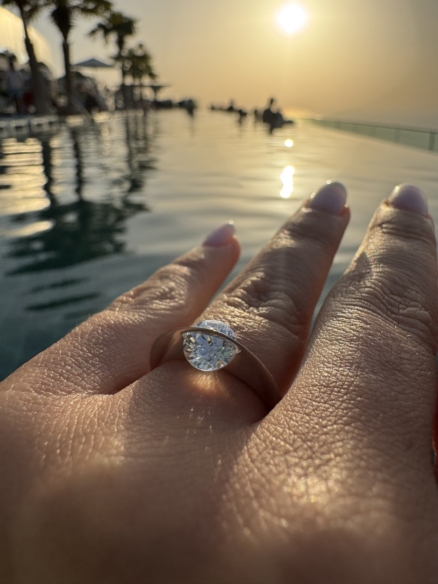 Anello Giroluce sul dito indossato con diamante che ruota, riflesso di luce al tramonto sull’acqua, atmosfera romantica e lussuosa.