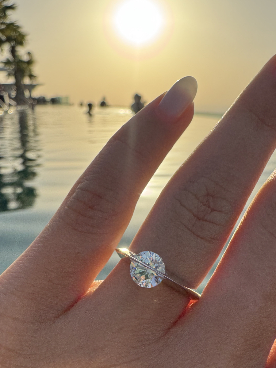 Mano con anello solitario Giroluce di Torinogioielli, con diamante rotante, fotografato al tramonto su uno sfondo di piscina a sfioro.